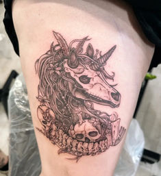 Black-and-grey thigh tattoo of a macabre unicorn skull with flowing mane, flowers and small human skulls woven into a spinal vertebra border, detailed linework fantasy-horror design.