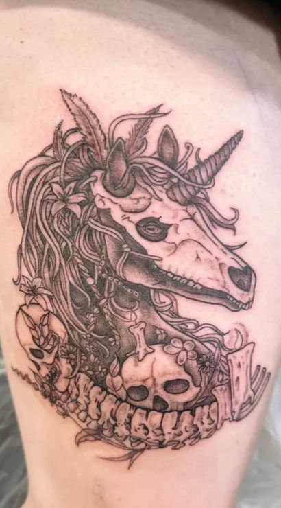 Black-and-grey thigh tattoo of a macabre unicorn skull with flowing mane, flowers and small human skulls woven into a spinal vertebra border, detailed linework fantasy-horror design.