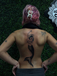Back view of a person with pink hair in a white clip displaying a large black mermaid tattoo down the spine, with smaller arm tattoos visible, posed against a green faux boxwood wall in a tattoo studio setting.