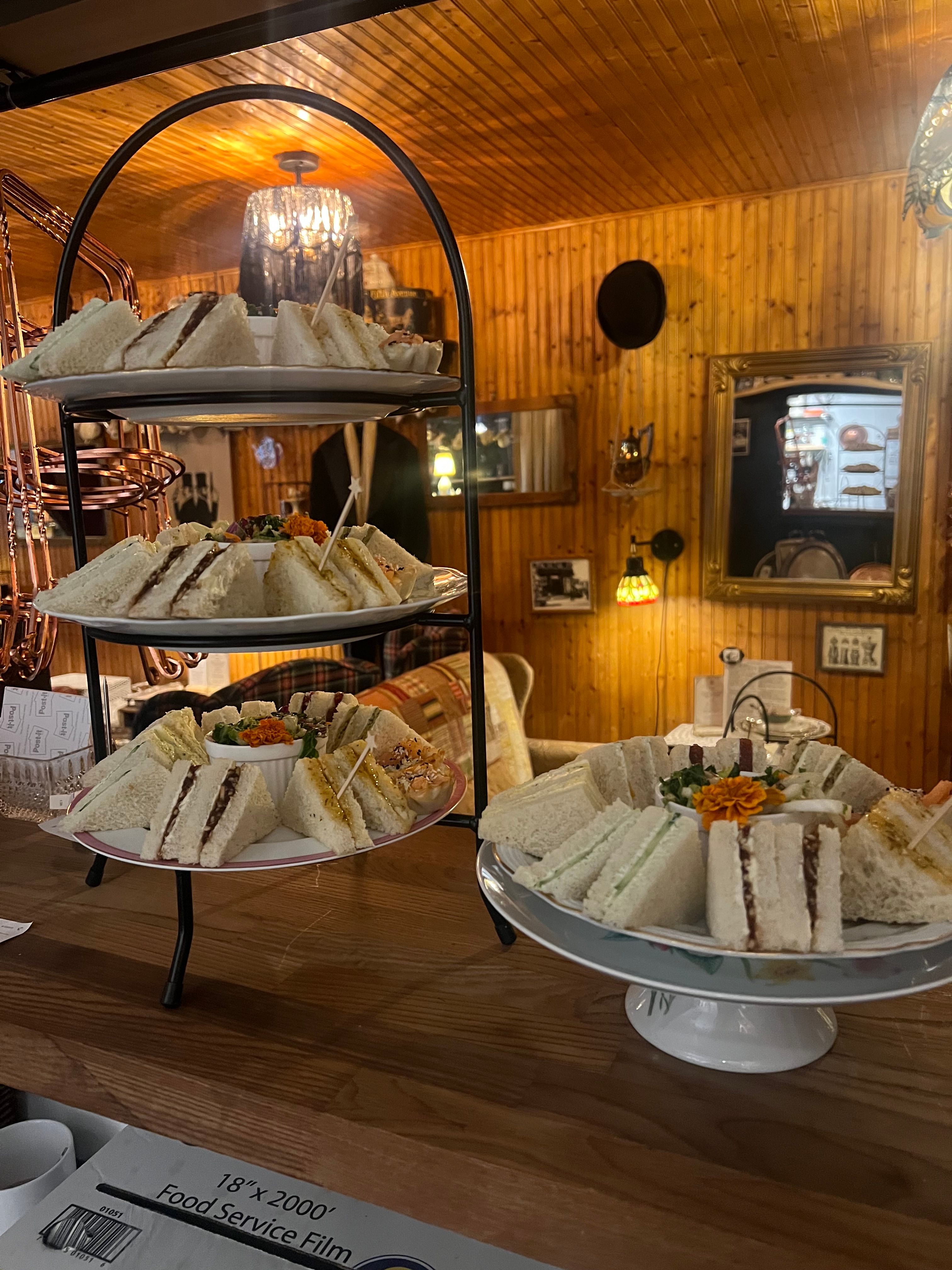 Tiered plates of crustless finger sandwiches and garnishes arranged for afternoon tea in a cozy wood‑paneled vintage tearoom with warm pendant lighting, mirrors and eclectic decor.