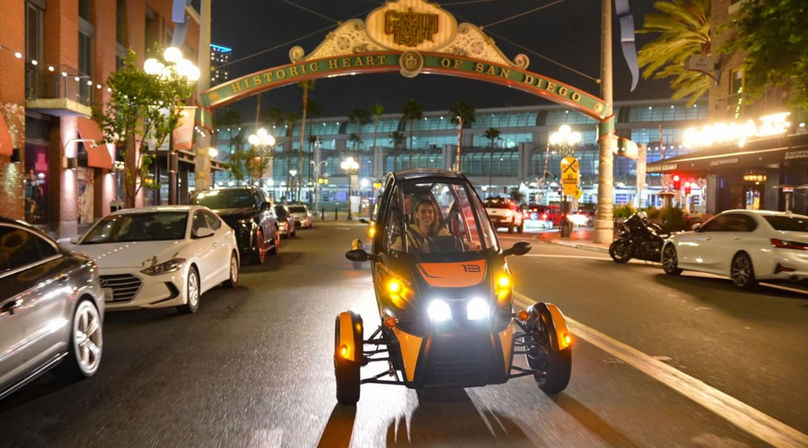 Orange three-wheeled electric vehicle with bright headlights driven under the Gaslamp Quarter historic arch in downtown San Diego at night, city street lined with parked cars, palm trees, and glowing street lamps.
