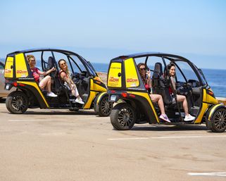 Four women smiling in two yellow open-sided electric tour cars parked at a sunny oceanfront overlook with clear blue sea and sky — scenic coastal tour.