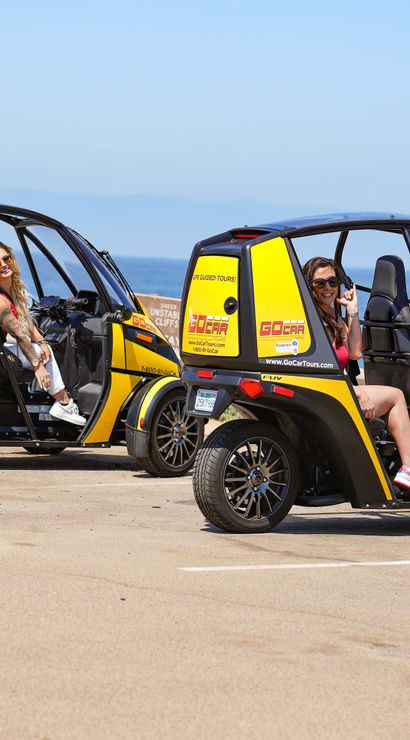 Four women seated in two bright yellow open-sided three-wheeled tour carts parked at an oceanfront viewpoint with the blue sea and clear sky in the background.