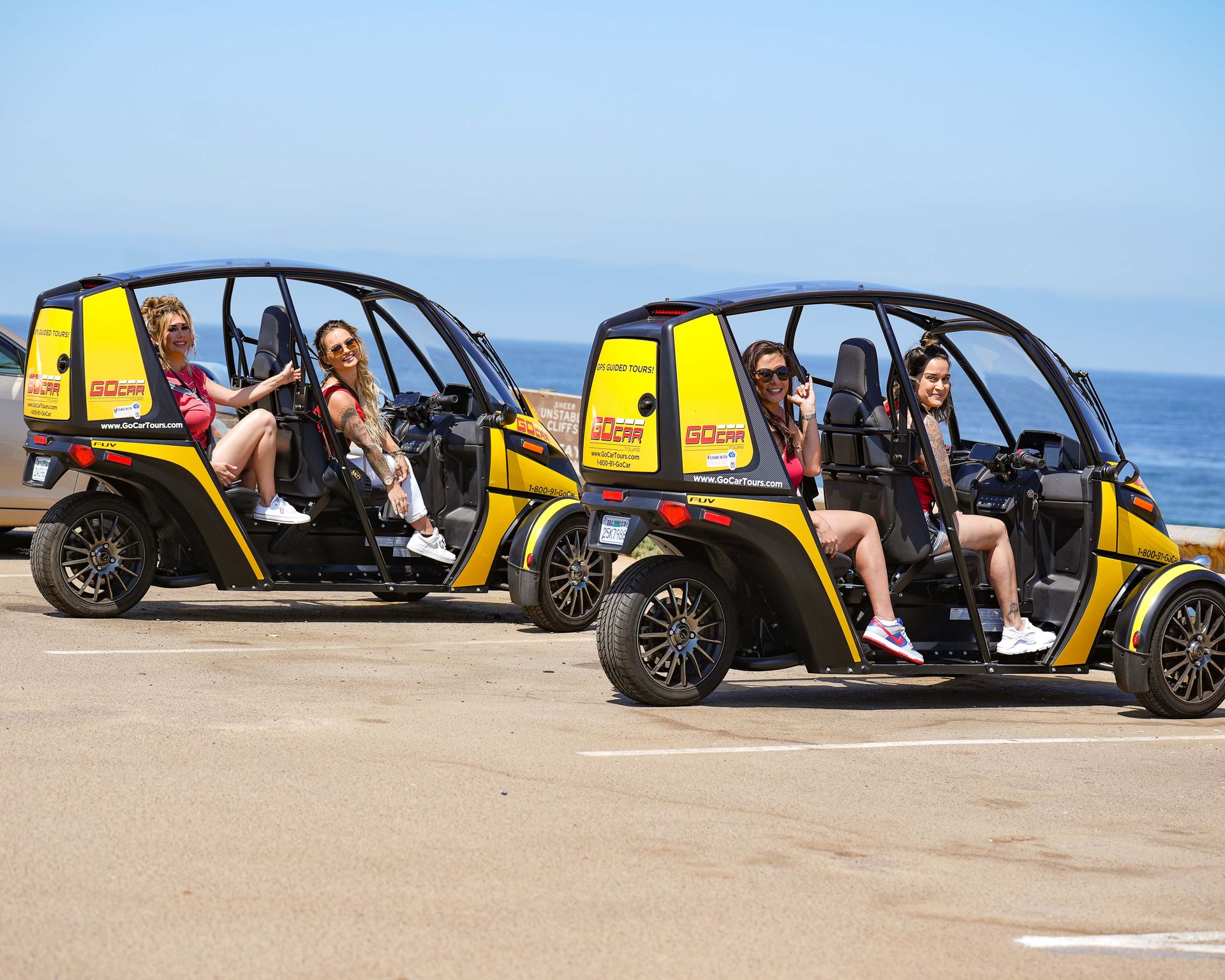 Four women seated in two bright yellow open-sided three-wheeled tour carts parked at an oceanfront viewpoint with the blue sea and clear sky in the background.