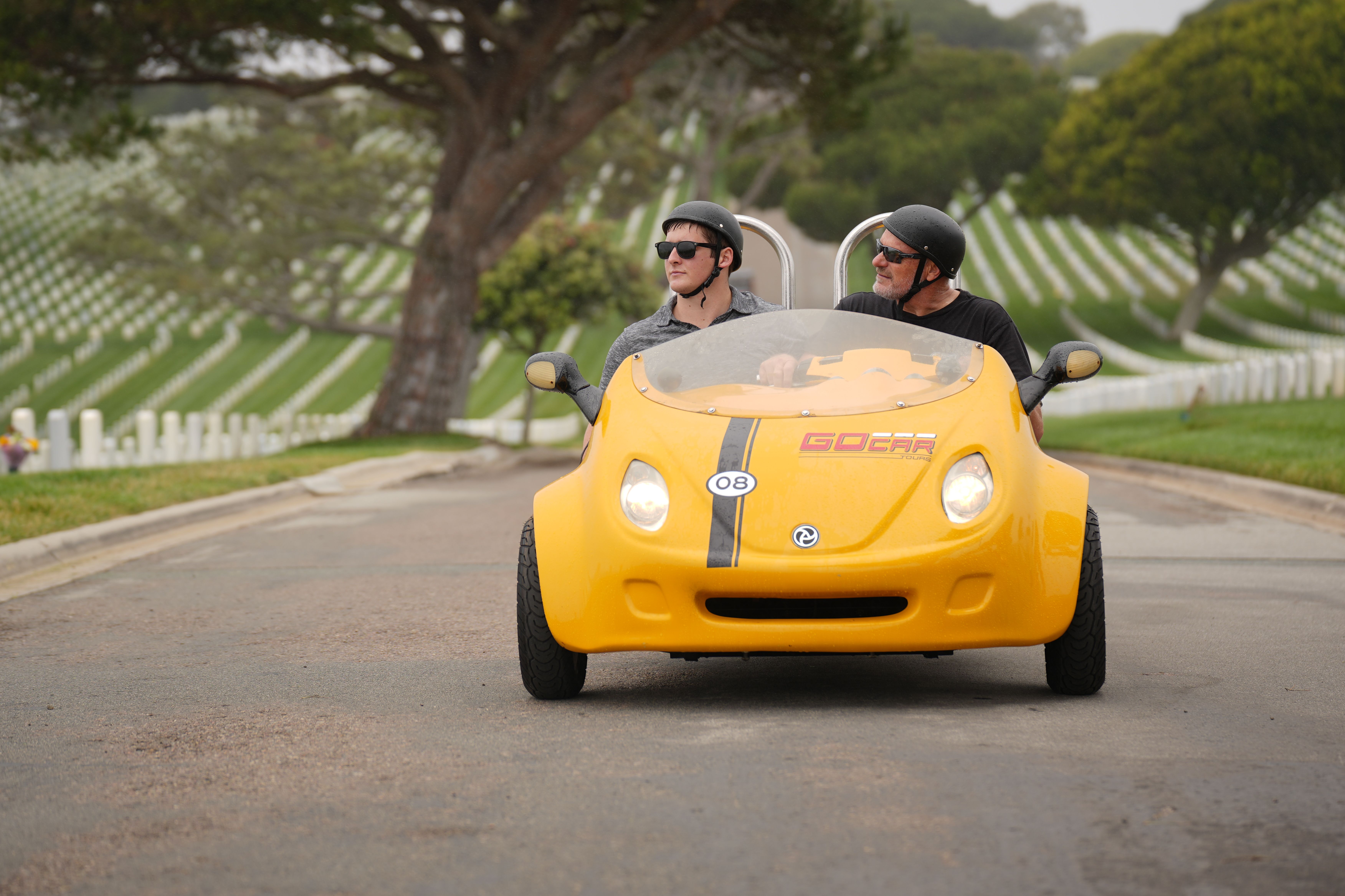 Two helmeted riders in a bright yellow two-seat tour car driving down a road through a hillside cemetery with neat rows of white headstones and trees