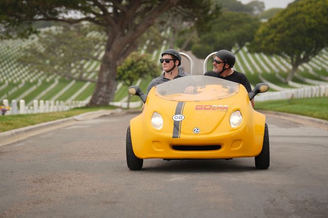 Two helmeted riders in a bright yellow two-seat tour car driving down a road through a hillside cemetery with neat rows of white headstones and trees