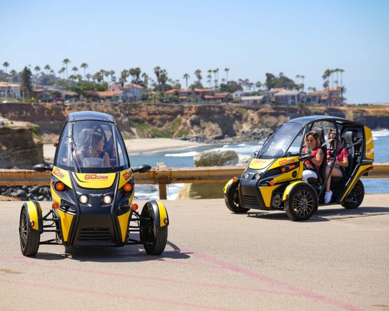Two yellow three-wheeled tour cars with passengers driving along a sunny coastal cliff road, palm trees and beachfront houses in the background