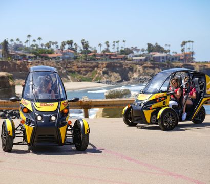 Two yellow three-wheeled tour cars with passengers driving along a sunny coastal cliff road, palm trees and beachfront houses in the background