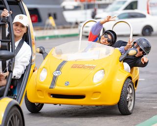 Smiling riders in a bright yellow two-seat open-top tour microcar wearing helmets and flashing peace signs on a fun urban sightseeing drive.