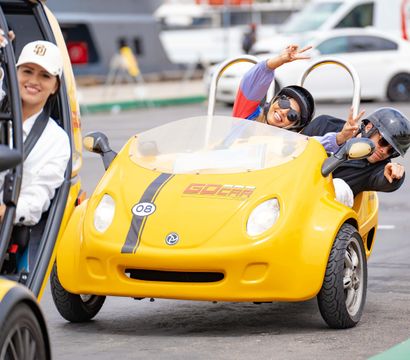 Smiling riders in a bright yellow two-seat open-top tour microcar wearing helmets and flashing peace signs on a fun urban sightseeing drive.