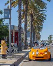 Bright yellow two-seat rental buggy with two helmeted tourists driving down a sunny, palm-lined coastal street under a clear blue sky