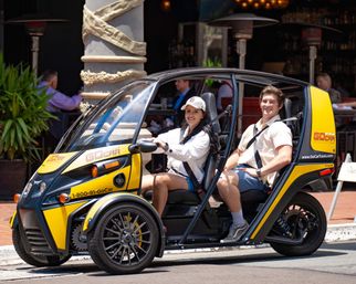 Smiling man and woman seated in a bright yellow two-seat open-sided sightseeing microcar on a sunny downtown street outside a café