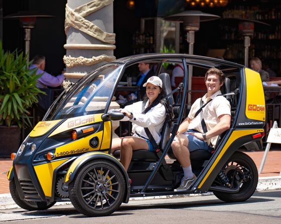 Smiling man and woman seated in a bright yellow two-seat open-sided sightseeing microcar on a sunny downtown street outside a café