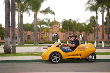 Two people wearing helmets smile in a yellow three-wheeled tour car parked along a palm-lined plaza with Spanish-style buildings and fountains in the background.