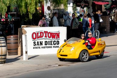 Couple smiling in a bright yellow two-seat tour car passing a state historic park sign in San Diego, busy pedestrian street scene