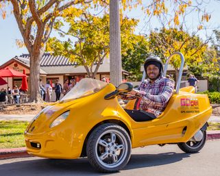 Person in a helmet and plaid shirt driving a bright yellow three-wheeled GPS-guided tour vehicle on a sunny, tree-lined street with autumn leaves and an outdoor café in the background.