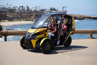 Two women in a compact yellow electric tour vehicle on a sunny oceanfront cliff road with sandy beach and palm trees.