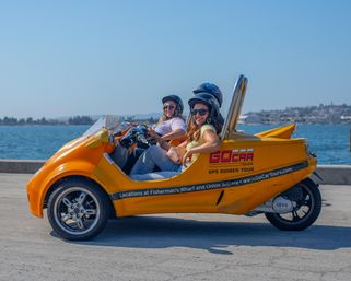 Two people wearing helmets and sunglasses riding a bright yellow three-wheeled sightseeing vehicle along a sunny waterfront promenade with blue bay water and a distant shoreline.