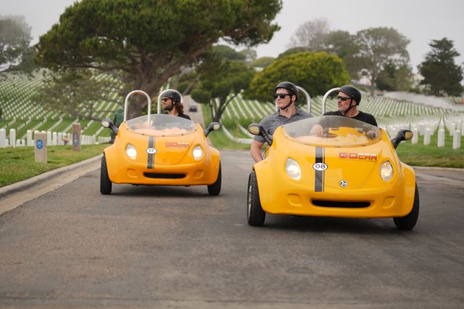 Two bright yellow three-wheeled tour cars with helmeted riders driving down a road through a cemetery of neatly aligned white headstones and green trees under an overcast sky.