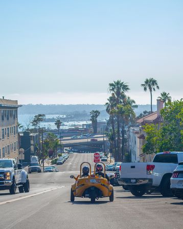 Sunny coastal city street descending toward a harbor and ocean, palm trees lining the block and parked trucks along the curb, with a bright yellow three-wheeled car carrying two helmeted riders driving downhill.