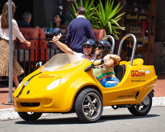 Smiling couple in helmets taking a selfie in a bright yellow two-seat sightseeing tour vehicle on a sunny downtown street with outdoor cafés
