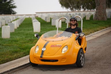 Person wearing a helmet and sunglasses driving a bright orange two-seat microcar along a road beside neatly lined white cemetery headstones on a green lawn.