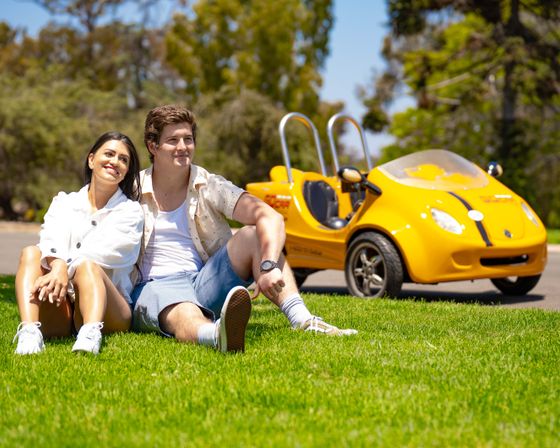 Smiling couple relaxing on a green park lawn on a sunny day with a bright yellow compact two-seat microcar parked on the path behind them