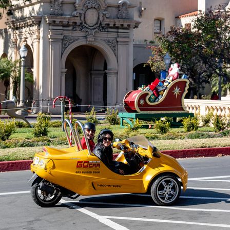 Two helmeted riders in a bright yellow three-wheeled tour car on a city street, passing a festive Santa sleigh display in front of ornate Spanish-style architecture.