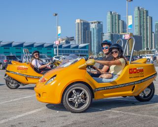 Two people wearing helmets and sunglasses riding bright yellow three-wheeled sightseeing vehicles in a sunny waterfront parking lot with a downtown high-rise skyline.