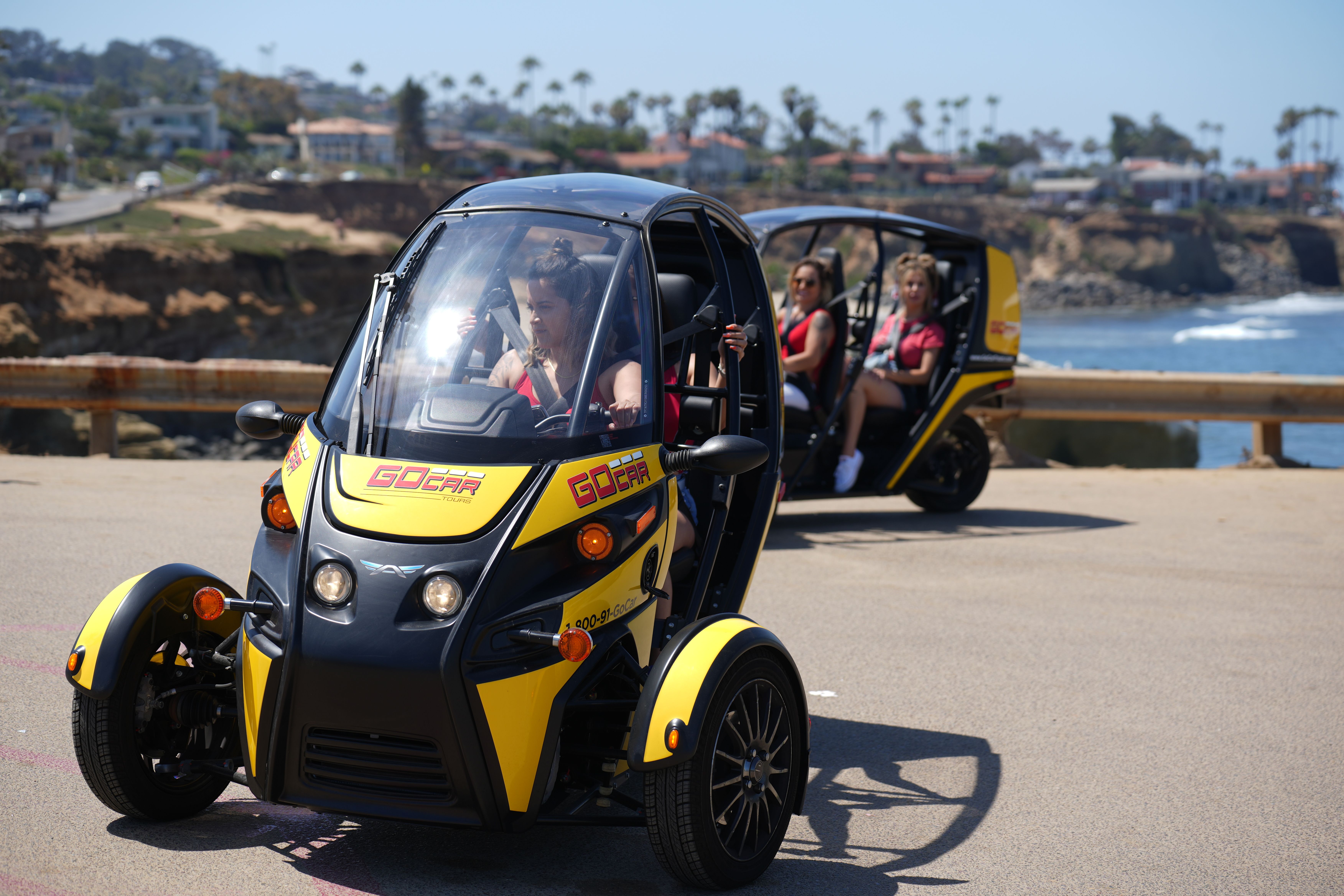 Bright sunny oceanfront scene of zippy yellow three-wheeled electric tour vehicles on a coastal cliff road, people buckled in, palm trees and rocky shoreline in the background.