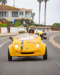 Driver wearing helmet and sunglasses cruising a bright yellow two-seat open-top tour car on a coastal road with palm trees and beachfront homes, a matching car following behind.