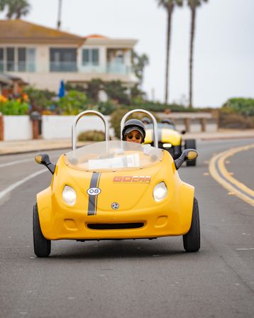 Driver wearing helmet and sunglasses cruising a bright yellow two-seat open-top tour car on a coastal road with palm trees and beachfront homes, a matching car following behind.