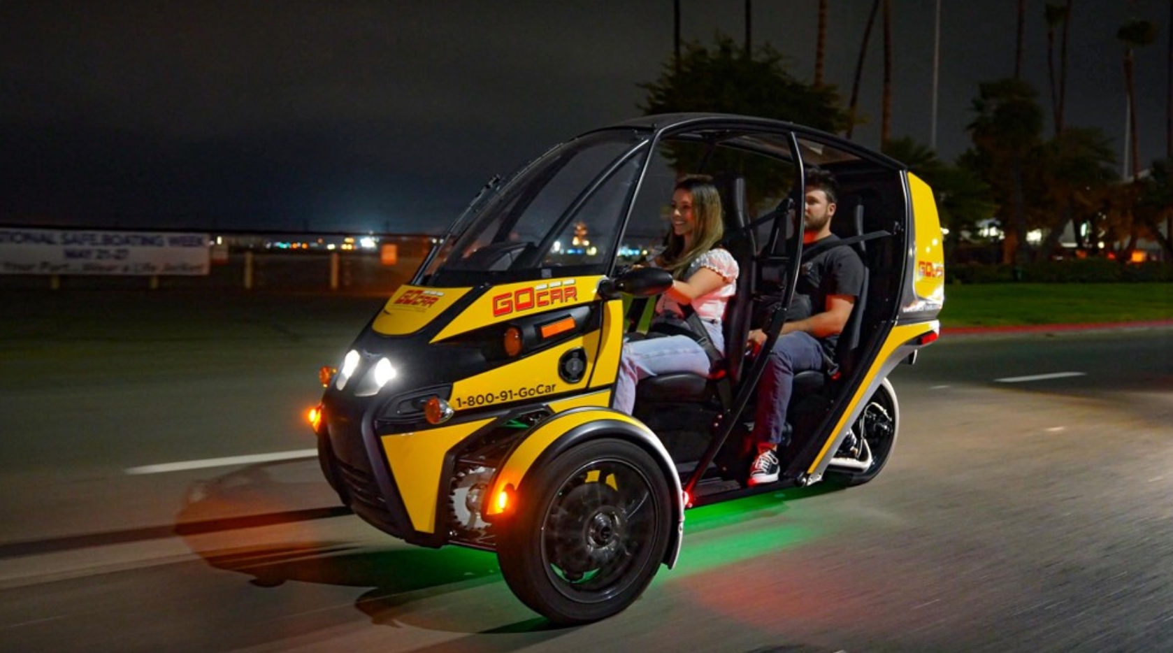 Yellow three-wheeled electric rental vehicle with two passengers driving at night along a palm-lined waterfront road, green underglow lights and headlights on
