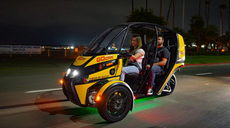 Yellow three-wheeled electric rental vehicle with two passengers driving at night along a palm-lined waterfront road, green underglow lights and headlights on