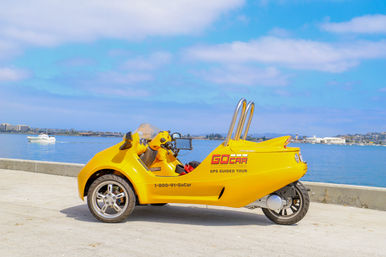 Bright yellow three-wheeled tour vehicle parked on a sunny seaside promenade with calm blue harbor, a white boat, and distant city skyline.