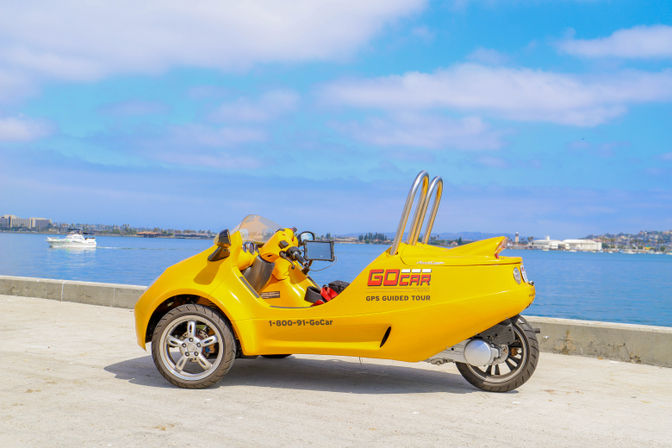 Bright yellow three-wheeled tour vehicle parked on a sunny seaside promenade with calm blue harbor, a white boat, and distant city skyline.