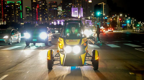 Yellow three-wheeled electric vehicle with bright twin headlights driven through a busy downtown street at night, city skyline and traffic lights glowing in the background