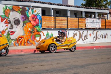Bright yellow three-wheeled tour vehicle with two helmeted riders on a sunny downtown street in front of a playful mural of a floral sugar-skull woman and an orange octopus.