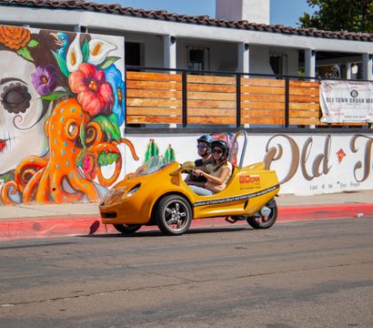 Bright yellow three-wheeled tour vehicle with two helmeted riders on a sunny downtown street in front of a playful mural of a floral sugar-skull woman and an orange octopus.