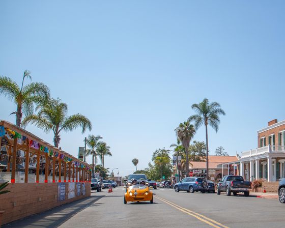 Vintage orange convertible on a sunny coastal California main street lined with palm trees, outdoor patios with colorful bunting and parked cars under a clear blue sky