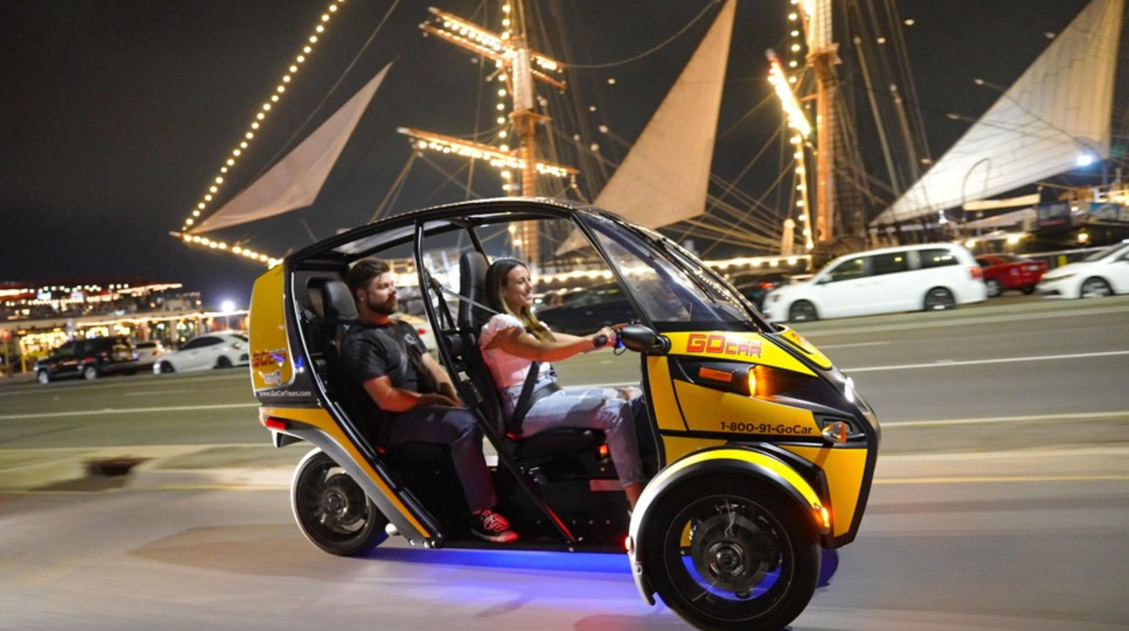 Yellow three-wheeled tour vehicle with two passengers cruising a lively waterfront at night past an illuminated tall ship and traffic.