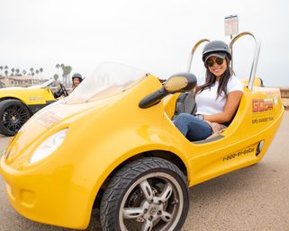 Smiling woman in helmet and sunglasses seated in a bright yellow two-seat rental tour car on a coastal boardwalk with palm trees and another yellow car in the background.