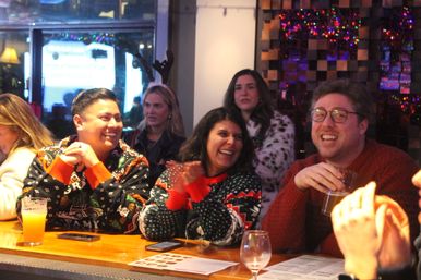 Group of friends in festive holiday sweaters laughing and clapping at a cozy bar, holding beers and cocktails under colorful holiday lights.