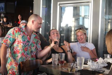 Friends at a lively brewery taproom: a man in a colorful Hawaiian shirt holds a microphone for a woman singing while another friend laughs at a table with drinks and a basket of snacks.