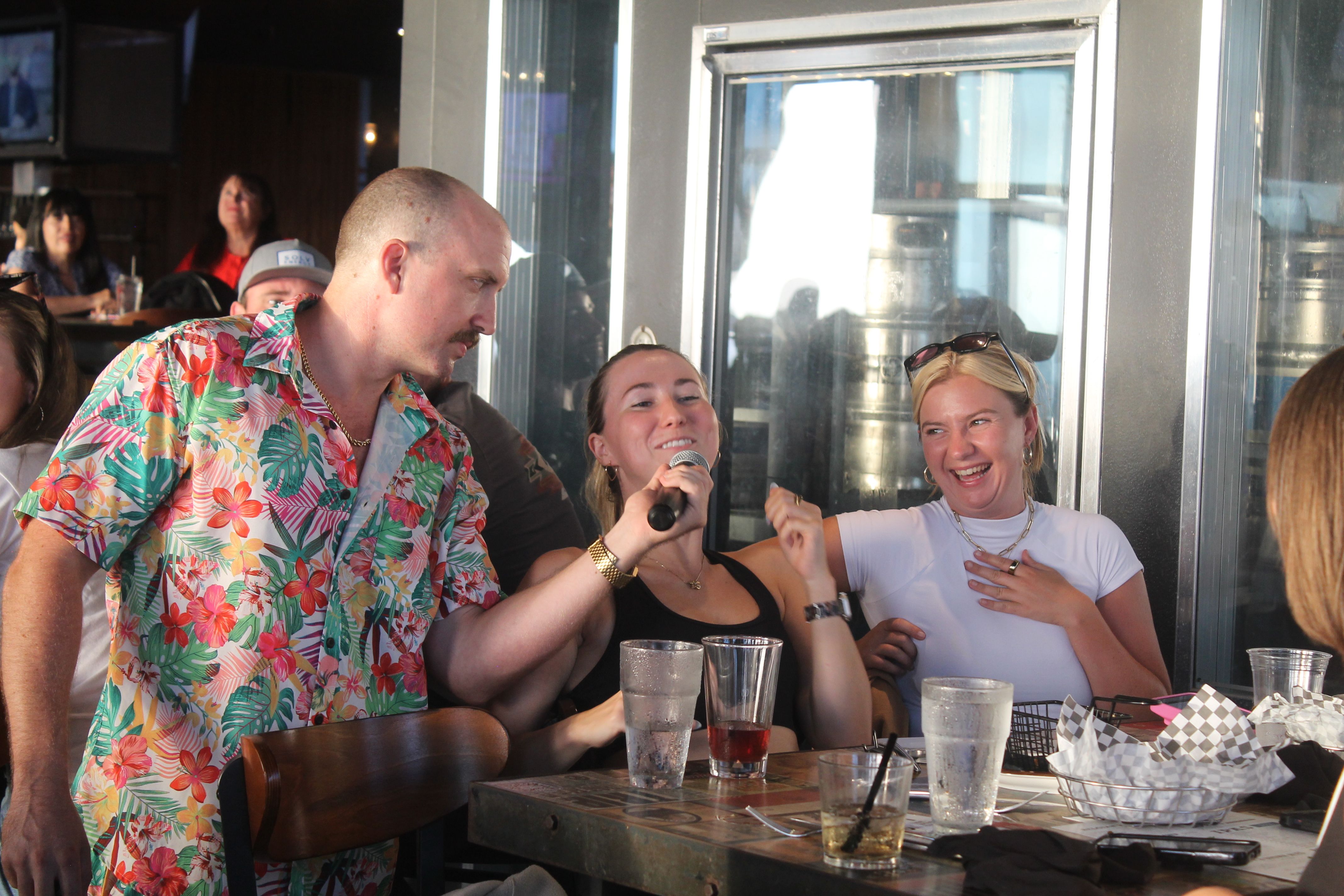 Man in bright floral shirt holding a microphone to a smiling woman while friends laugh at a sunlit bar patio table with drinks — lively karaoke singalong.