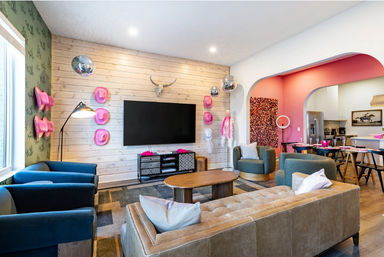 Bright open-concept lounge with a wood-plank TV wall and mounted flat-screen, playful pink cowboy hats and disco balls, decorative steer skull, teal armchairs and tan tufted sofa around a wooden coffee table, arched doorway to a pink-accented dining area and kitchen.