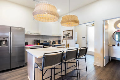 Bright modern kitchen with white island and sink, three gray bar stools, woven rattan pendant lights, stainless steel refrigerator and oven, open layout with wood-look flooring.