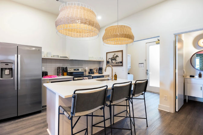 Bright modern kitchen with white island and sink, three gray bar stools, woven rattan pendant lights, stainless steel refrigerator and oven, open layout with wood-look flooring.