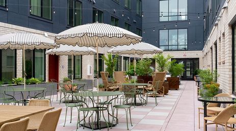 Cheerful urban courtyard patio with striped umbrellas, green bistro tables, wicker chairs, potted plants and overhead string lights between modern brick-and-glass buildings.