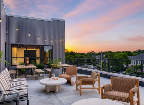 Urban rooftop terrace at sunset with woven lounge chairs, striped sofa, round coffee tables, picnic-style dining bench, string lights and potted plants overlooking a tree-lined neighborhood.